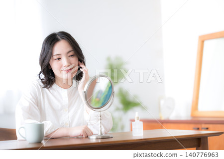 一位微笑的女士對著鏡子,正在到臉頰和皮膚上塗抹保養品。美麗的肌膚和自我照顧。 一位微笑的女士對著鏡子,正在到臉頰和皮膚上塗抹保養品。美麗的肌膚和自我照顧。 140776963