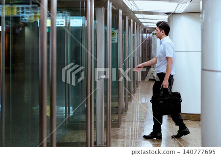 A Japanese office worker in a short-sleeved shirt, carrying a business bag, walks through the glass entrance of an underground passageway during his commute or travel. A Japanese office worker in a short-sleeved shirt, carrying a business bag, walks through the glass entrance of an underground passageway during his commute or travel. 140777695