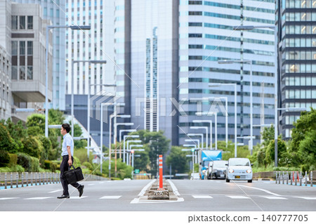 A Japanese male office worker in his 30s, wearing a short-sleeved shirt and carrying a bag, walks across a pedestrian crossing in a city lined with skyscrapers on his way to work. A Japanese male office worker in his 30s, wearing a short-sleeved shirt and carrying a bag, walks across a pedestrian crossing in a city lined with skyscrapers on his way to work. 140777705