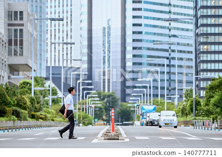 A Japanese man in his 30s, wearing a short-sleeved shirt and working for a company, walks across a pedestrian crossing on a city street, with a backdrop of skyscrapers. A Japanese man in his 30s, wearing a short-sleeved shirt and working for a company, walks across a pedestrian crossing on a city street, with a backdrop of skyscrapers. 140777711