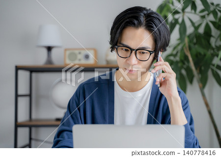 A man at home is talking on his smartphone while looking at his computer. A man at home is talking on his smartphone while looking at his computer. 140778416