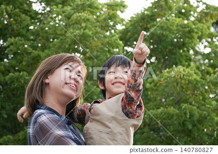 Happy Asian mother holding her little son as he points up to the sky outdoors with green trees background. Happy Asian mother holding her little son as he points up to the sky outdoors with green trees background. 140780827