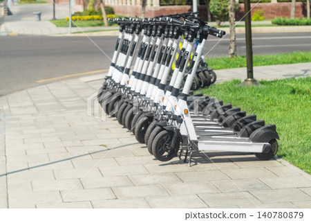 Rows of parked electric scooters for customer rental on the edge of a pavement lined with cobblestones in the city center. Regulation of the movement of private transport of urban residents Rows of parked electric scooters for customer rental on the edge of a pavement lined with cobblestones in the city center. Regulation of the movement of private transport of urban residents 140780879