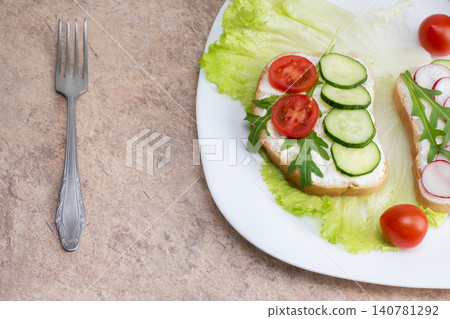 Sandwich with cottage cheese, cucumbers and cherry tomatoes and radish on a white plate on the table. Plate with a fork on the table with a white plate with a salad of vegetables. Sandwich with cottage cheese, cucumbers and cherry tomatoes and radish on a white plate on the table. Plate with a fork on the table with a white plate with a salad of vegetables. 140781292