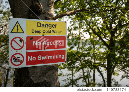 Danger sign warning of deep cold water. No access. No swimming. Warning sign on a tree with trees and lake behind. Danger sign warning of deep cold water. No access. No swimming. Warning sign on a tree with trees and lake behind. 140782374