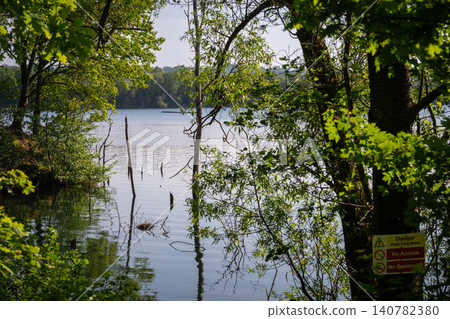 Mercers Lake near Merstham, Surrey, UK. View through trees to the cool waters of the lake. A danger sign is on a tree. Mercers Lake near Merstham, Surrey, UK. View through trees to the cool waters of the lake. A danger sign is on a tree. 140782380