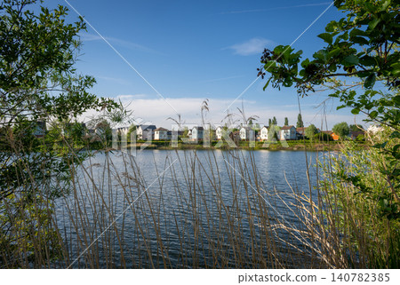 A modern housing development next to the Holmethorpe Lagoons in Mersham, Surrey, UK. Trees and reeds in the foreground. Landscape orientation. Focus on foreground. 140782385