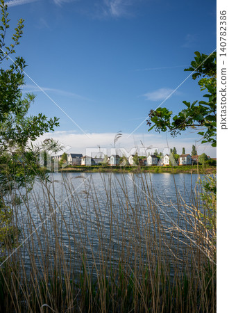 A modern housing development next to the Holmethorpe Lagoons in Mersham, Surrey, UK. Trees and reeds in the foreground. Portrait orientation. A modern housing development next to the Holmethorpe Lagoons in Mersham, Surrey, UK. Trees and reeds in the foreground. Portrait orientation. 140782386