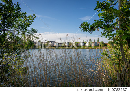 A modern housing development next to the Holmethorpe Lagoons in Mersham, Surrey, UK. Trees and reeds in the foreground. Landscape orientation. A modern housing development next to the Holmethorpe Lagoons in Mersham, Surrey, UK. Trees and reeds in the foreground. Landscape orientation. 140782387