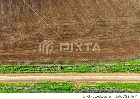 Aerial downward view of a green grass lined dirt road leading through cultivated agricultural fields, showcasing rural farmland patterns and textures beneath soft natural light in NSW, Australia. Aerial downward view of a green grass lined dirt road leading through cultivated agricultural fields, showcasing rural farmland patterns and textures beneath soft natural light in NSW, Australia. 140782467