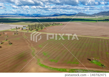 Aerial view of cultivated agricultural fields surrounded by lush green landscape, with a dirt road leading through the farmland, highlighting rural patterns and textures in NSW, Australia. Aerial view of cultivated agricultural fields surrounded by lush green landscape, with a dirt road leading through the farmland, highlighting rural patterns and textures in NSW, Australia. 140782471