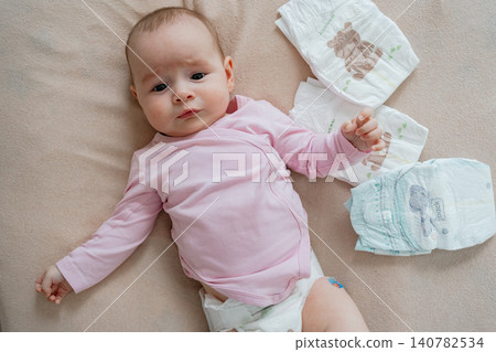 Close-up overhead shot of a sweet baby girl in pink clothes lying down on a soft blanket, surrounded by clean diapers during morning care. Adorable newborn infant lying on a neutral bed sheet with Close-up overhead shot of a sweet baby girl in pink clothes lying down on a soft blanket, surrounded by clean diapers during morning care. Adorable newborn infant lying on a neutral bed sheet with 140782534