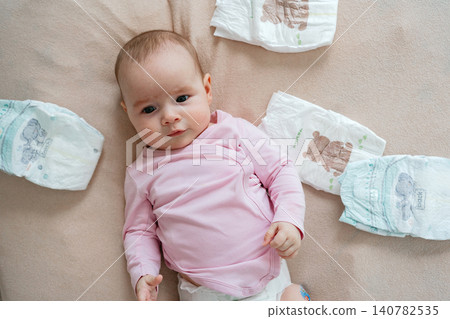 Calm newborn baby in a pink long sleeve bodysuit lying on a soft blanket next to folded clean diapers, looking up curiously. Top view. High angle portrait of a cute infant girl during her morning Calm newborn baby in a pink long sleeve bodysuit lying on a soft blanket next to folded clean diapers, looking up curiously. Top view. High angle portrait of a cute infant girl during her morning 140782535