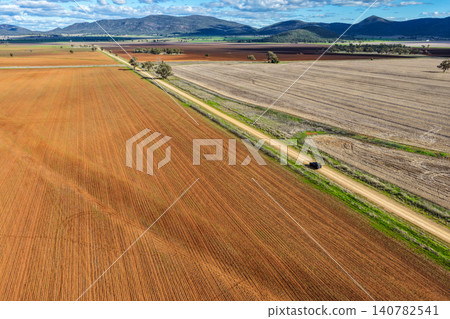 Aerial view of cultivated agricultural fields surrounded by lush green landscape, with a car travelling through dirt road leading through the farmland, highlighting rural patterns and textures. Aerial view of cultivated agricultural fields surrounded by lush green landscape, with a car travelling through dirt road leading through the farmland, highlighting rural patterns and textures. 140782541