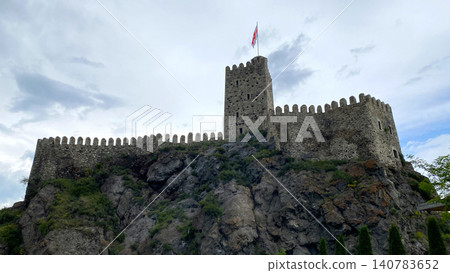 Rabat Fortress towering above rocky cliffs. Low angle view of medieval walls and watchtower crowned with Georgian flag Rabat Fortress towering above rocky cliffs. Low angle view of medieval walls and watchtower crowned with Georgian flag 140783652