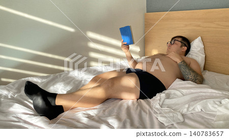 Relaxed man reading a book while lying on a bed in a bright bedroom filled with natural morning sunlight 140783657
