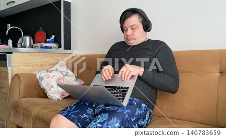 Funny man wearing wireless headphones and using a laptop while sitting on a sofa at home. Humorous facial expression Funny man wearing wireless headphones and using a laptop while sitting on a sofa at home. Humorous facial expression 140783659