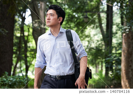 A Japanese man in his 30s, working in sales, walks through a lush green park on a hot summer day, sweating as he walks, either commuting or on his way to work. A Japanese man in his 30s, working in sales, walks through a lush green park on a hot summer day, sweating as he walks, either commuting or on his way to work. 140784116