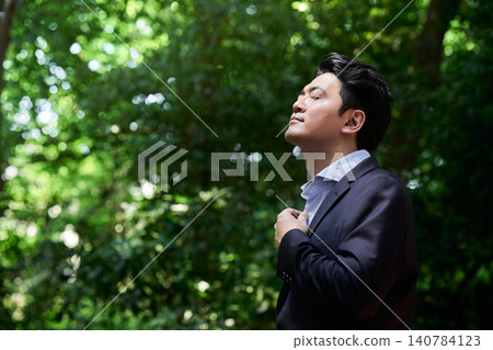 一位三十多歲的男子，身穿西裝，閉上眼睛，深吸一口氣，在戶外清新的綠蔭中放鬆身心，感受著空氣的涼爽。 140784123