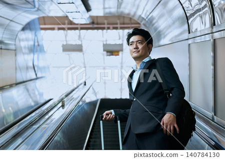 A Japanese male office worker in his 30s riding an escalator with a carry-on bag while traveling for a business trip. A Japanese male office worker in his 30s riding an escalator with a carry-on bag while traveling for a business trip. 140784130