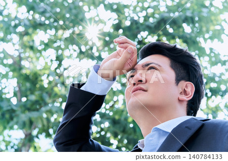 A Japanese office worker in his 30s, looking unwell and on the verge of heatstroke, shields his eyes from the sun with his hands in the shade under a tree outdoors. A Japanese office worker in his 30s, looking unwell and on the verge of heatstroke, shields his eyes from the sun with his hands in the shade under a tree outdoors. 140784133