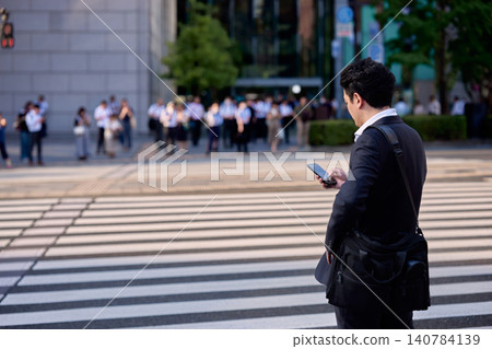 一位日本商人在高峰時段穿過城市辦公區的人行橫道時正在使用智慧型手機。 一位日本商人在高峰時段穿過城市辦公區的人行橫道時正在使用智慧型手機。 140784139