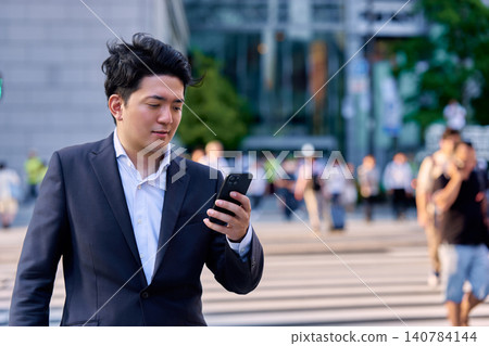 傍晚時分,在繁華的市中心辦公區,一位商人正在熙熙攘攘的十字路口使用智慧型手機。 傍晚時分,在繁華的市中心辦公區,一位商人正在熙熙攘攘的十字路口使用智慧型手機。 140784144