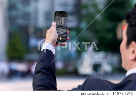 A faceless Japanese office worker in a suit is seen taking pictures of a city streetscape with his smartphone. A faceless Japanese office worker in a suit is seen taking pictures of a city streetscape with his smartphone. 140784146