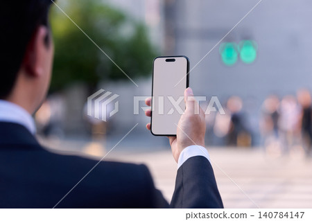 The back view and hands of a faceless office worker in a city office district, holding a smartphone and looking at the screen while on his way home. The back view and hands of a faceless office worker in a city office district, holding a smartphone and looking at the screen while on his way home. 140784147