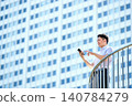 A Japanese businessman in his 30s, wearing a short-sleeved shirt, is operating his smartphone outdoors in an office building. 140784279