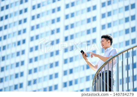 A Japanese businessman in his 30s, wearing a short-sleeved shirt, is operating his smartphone outdoors in an office building. A Japanese businessman in his 30s, wearing a short-sleeved shirt, is operating his smartphone outdoors in an office building. 140784279