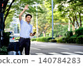 A male office worker with a suitcase is waiting for a taxi he booked using a ride-hailing app on his smartphone while on a business trip. 140784283