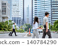 A businessman, pulling a carry-on bag, walks across a crosswalk in a city lined with skyscrapers, passing by people. 140784290