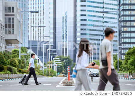一位商人牽手提行李,走過摩天大樓林立的城市的人行橫道,與行人擦肩而過。 一位商人牽手提行李,走過摩天大樓林立的城市的人行橫道,與行人擦肩而過。 140784290