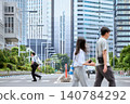 A Japanese businessman runs across a pedestrian crossing on a city street lined with skyscrapers, passing by people. 140784292