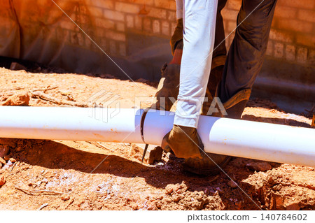 Worker holds white pipe and uses tool to cut it while on dirt surface at construction site. Worker holds white pipe and uses tool to cut it while on dirt surface at construction site. 140784602
