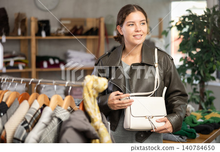 Sale of winter clothing collection. Positive young girl standing against background of shelves with clothes in modern store and choosing bag Sale of winter clothing collection. Positive young girl standing against background of shelves with clothes in modern store and choosing bag 140784650