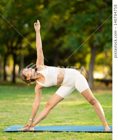 Girl in park on green grass of lawn performs Trikonasana, triangle asana Girl in park on green grass of lawn performs Trikonasana, triangle asana 140784758