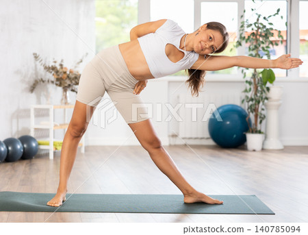 Young woman yoga trainer performing triangle pose on black rug Young woman yoga trainer performing triangle pose on black rug 140785094