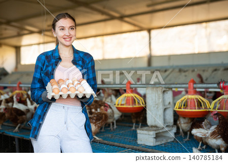 Smiling european woman in plaid shirt and cap collecting eggs in chicken farm 140785184