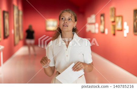 Young girl holding an information booklet looks at paintings in an art exhibition in museum 140785216