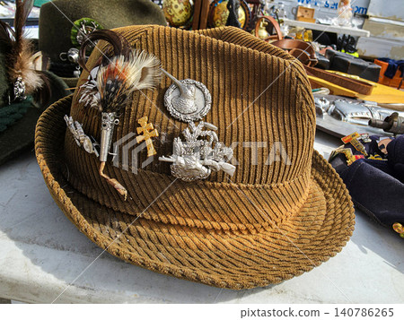 A brown corduroy fedora hat sits on a table at a flea market. The hat is decorated with pins, feathers, and a metal cross. Surrounding items hint at a rustic market atmosphere. 140786265