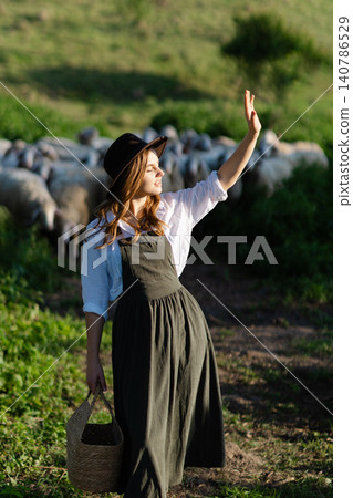 Shepherdess waving hand holding wicker basket walking on meadow with sheep flock in background Shepherdess waving hand holding wicker basket walking on meadow with sheep flock in background 140786529