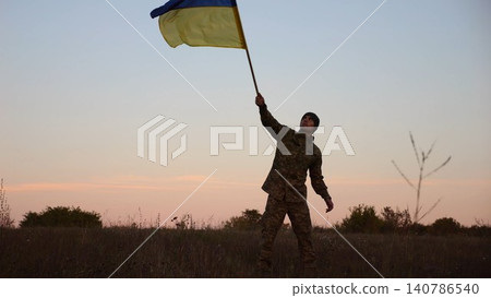 Male military waving blue-yellow banner on liberated territory occupied by russia. Young soldier of ukrainian army lifted flag of Ukraine in honor of victory against russian aggression at dusk 140786540