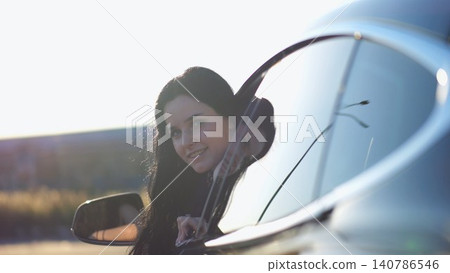 Young beautiful girl leaning out of driver window at modern electric car and looking into camera. Portrait of attractive smiling woman in alternative electrical vehicle. Slow motion Young beautiful girl leaning out of driver window at modern electric car and looking into camera. Portrait of attractive smiling woman in alternative electrical vehicle. Slow motion 140786546