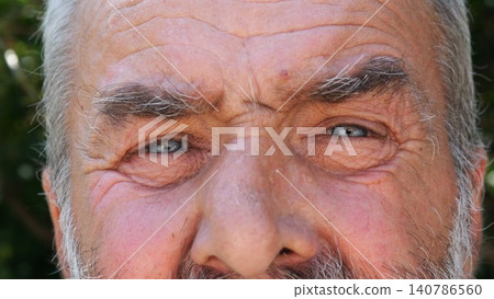 Blue eyes of elderly male blinking and looking into camera outdoor. Wrinkled face of senior human with a sad sight. Upset facial expression of pensioner. Close up Blue eyes of elderly male blinking and looking into camera outdoor. Wrinkled face of senior human with a sad sight. Upset facial expression of pensioner. Close up 140786560