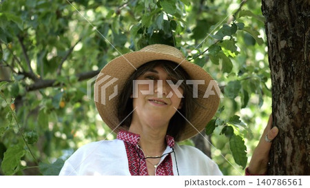 Portrait of ukrainian woman in straw hat against the background of summer garden. Adult smiling lady wearing an embroidered shirt looking into camera outdoor. National identity and patriotism concept Portrait of ukrainian woman in straw hat against the background of summer garden. Adult smiling lady wearing an embroidered shirt looking into camera outdoor. National identity and patriotism concept 140786561