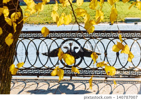 picturesque metal railings with a double-headed eagle and the crown of the Russian Empire through the yellow leaves on embankment in St. Petersburg, sunny autumn day, incredible sun glare Russia picturesque metal railings with a double-headed eagle and the crown of the Russian Empire through the yellow leaves on embankment in St. Petersburg, sunny autumn day, incredible sun glare Russia 140787007