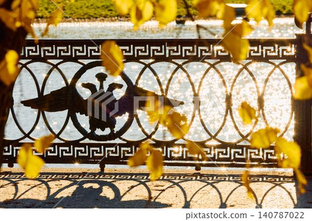 picturesque metal railings with a double-headed eagle and the crown of the Russian Empire through the yellow leaves on embankment in St. Petersburg, sunny autumn day, incredible sun glare Russia 140787022