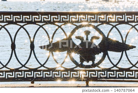picturesque metal railings with a double-headed eagle and the crown of the Russian Empire on embankment in St. Petersburg, Russia, walking people, sunny autumn day, incredible sun glare Russia 140787064